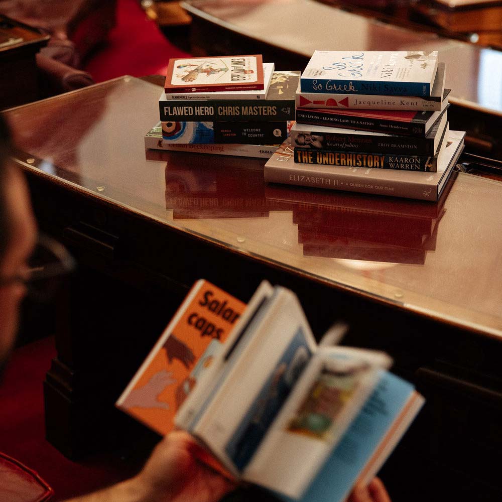 A person reads a book with a pile of additional books on a desk in front of them.