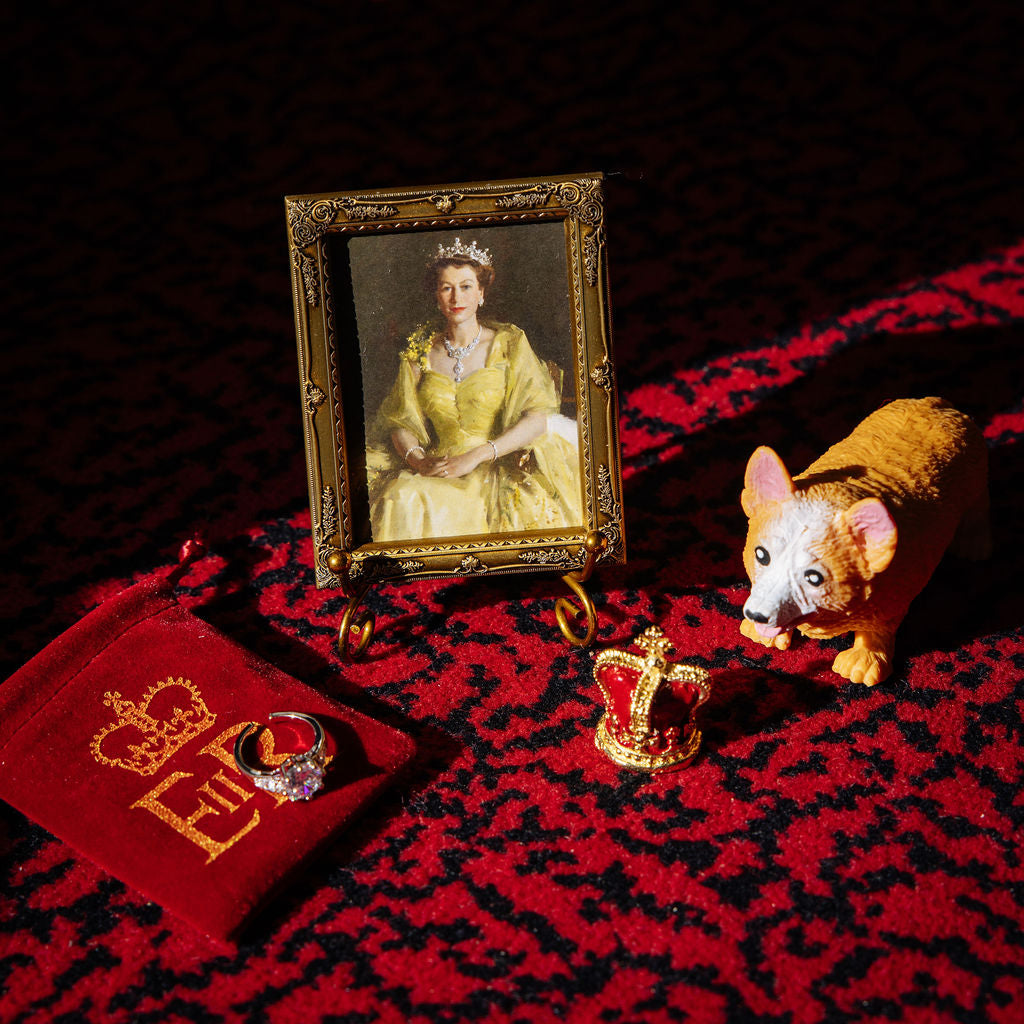 Royal-themed products arranged on red carpet, including a framed portrait of Queen Elizabeth II, a replica ring and crown and a corgi toy. 