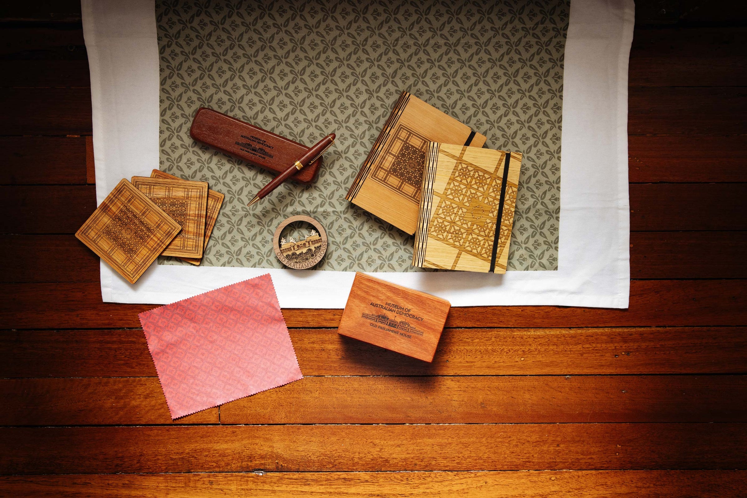 A group of products arranged on a wooden floor including notebooks, coasters, a pen and a tea towel. 