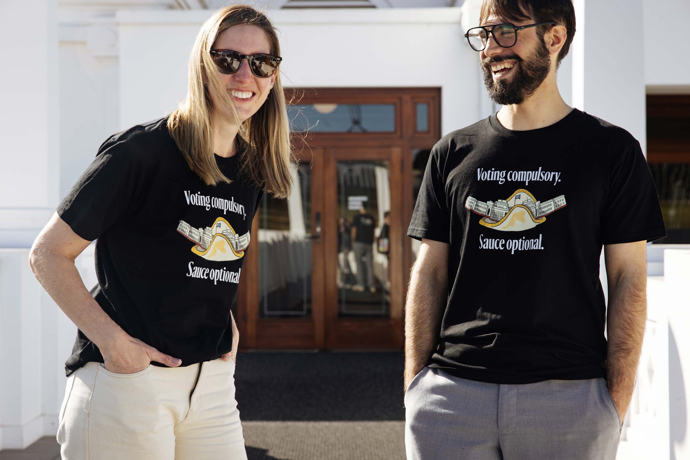 Two people wearing black t-shirts with the words 'Voting compulsory. Sauce Optional' around a sausage sandwich design. 