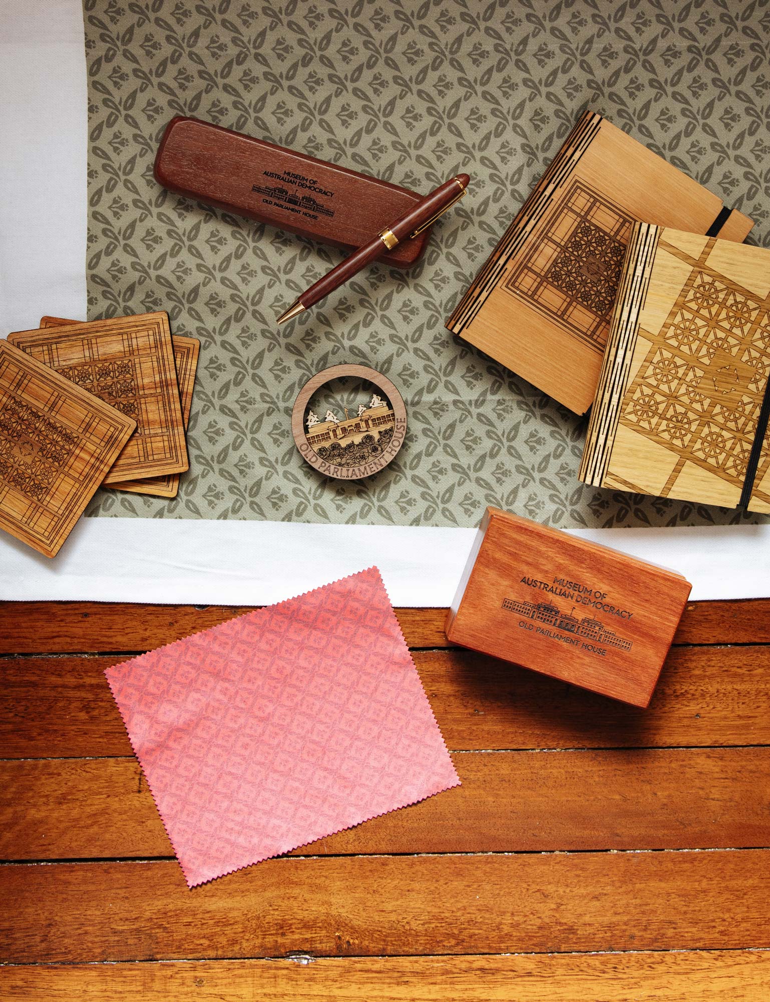 A group of products arranged on a wooden floor including notebooks, coasters, a pen and a tea towel. 